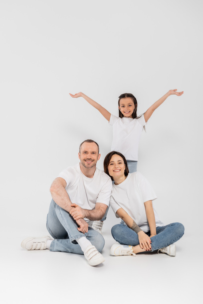 preteen daughter standing with outstretched hands near happy parents in white t-shirts and blue denim jeans looking at camera and posing together on grey background, child protection day  - Foto, afbeelding