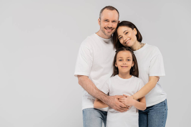 pais felizes abraçando filha alegre enquanto estão juntos em jeans jeans jeans azul e camisetas brancas e olhando para a câmera no fundo cinza, dia das crianças feliz, 1 de junho - Foto, Imagem
