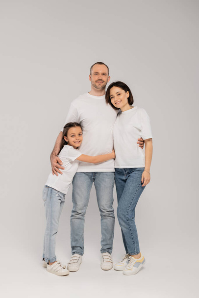 joyous preteen girl hugging father near mother with tattoo while standing together in blue denim jeans and white t-shirts and looking at camera on grey background, Happy children's day,  - Fotoğraf, Görsel