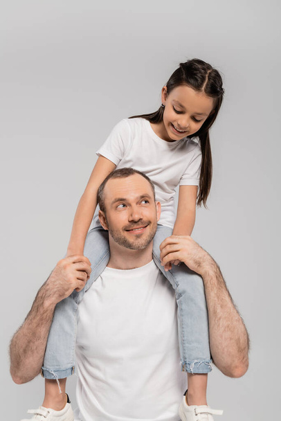 szczęśliwy nastoletnia córka z długimi brunetkami włosy siedzi na ramionach nieogolony i wesoły ojciec z włosia na szarym tle, dzień ochrony dziecka, ojciec i córka  - Zdjęcie, obraz