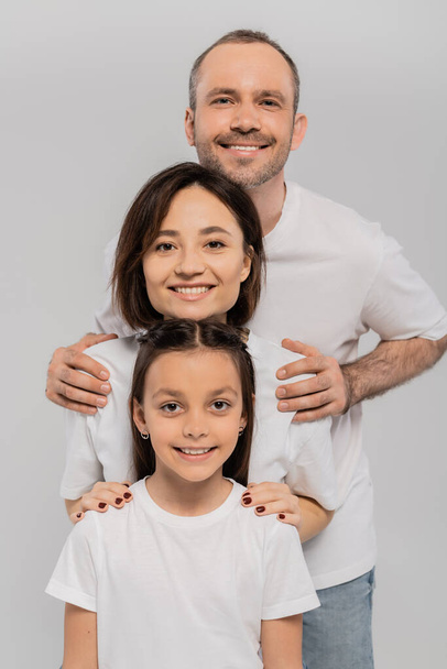boldog család fehér pólóban nézi kamera és pózol együtt szürke háttér, Nemzetközi gyermekvédelmi nap, pozitív szülők és lánya  - Fotó, kép