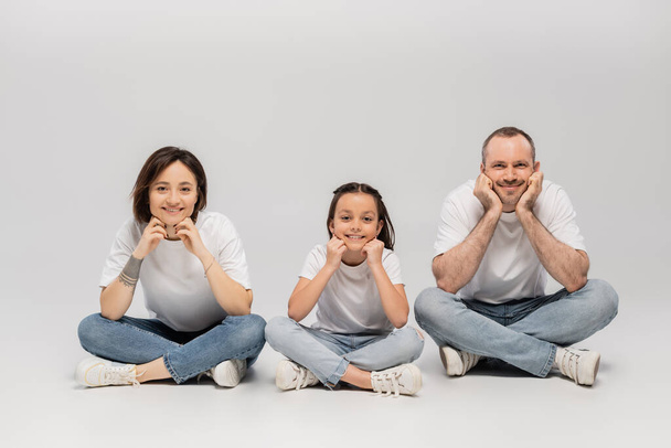 χαρούμενος πατέρας και μητέρα με τατουάζ με κοντά μαλλιά και χαρούμενη προεφηβική κόρη κάθεται με σταυρωμένα τα πόδια σε λευκό t-shirts και μπλε τζιν τζιν σε γκρι φόντο, ευτυχισμένη ημέρα των παιδιών  - Φωτογραφία, εικόνα
