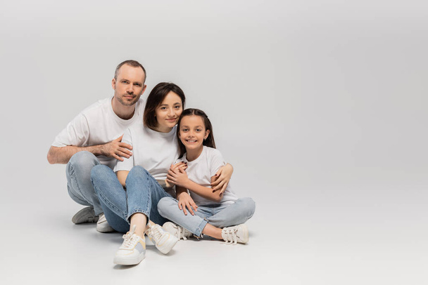 joyous father and mother with short hair hugging cheerful preteen daughter while sitting with crossed legs in white t-shirts and blue denim jeans on grey background, Happy children's day  - 写真・画像