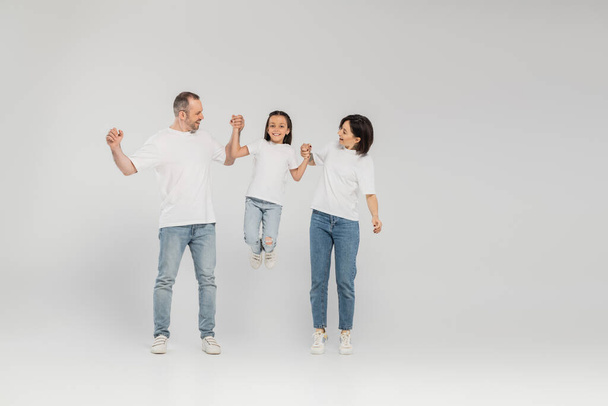 full length of happy parents in white t-shirts and blue denim jeans holding hands and lifting preteen daughter while standing together on grey background, International child protection day in June  - Фото, изображение