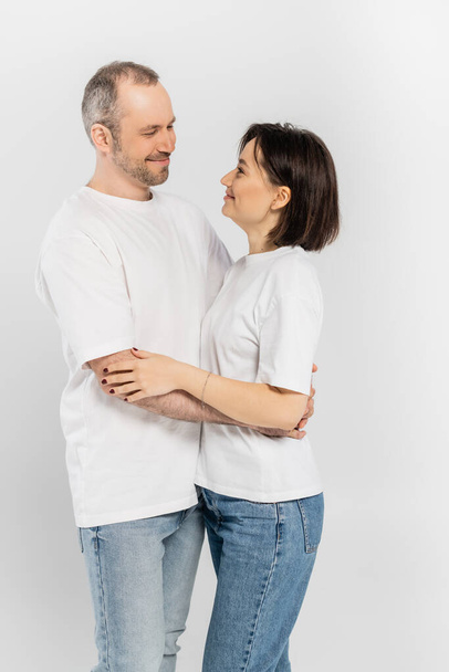 cheerful and tattooed woman with short brunette hair hugging joyous husband with bristle while standing together in white t-shirts and looking at each other isolated on grey background, happy couple  - Photo, Image