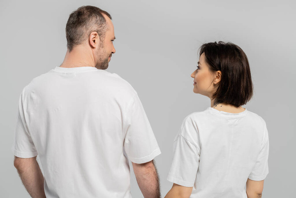Kısa esmer saçlı, neşeli ve dövmeli bir kadının beyaz tişörtlerle gri arka planda izole edilmiş mutlu çift tişörtleriyle kocasına baktığı arka plan.  - Fotoğraf, Görsel
