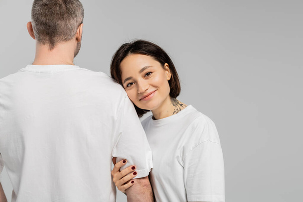 cheerful and tattooed woman with short brunette hair leaning on shoulder of husband while standing together in white t-shirts and looking at camera isolated on grey background, happy couple  - Фото, изображение