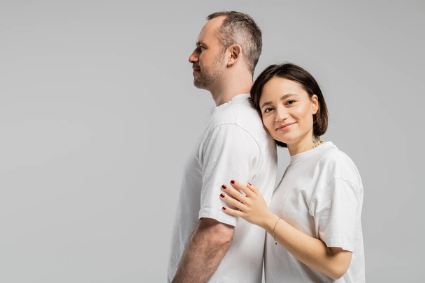 tattooed woman with short brunette hair leaning on back of husband while standing together in white t-shirts and looking at camera isolated on grey background, happy couple  - Фото, изображение