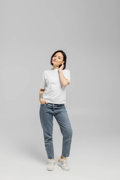full length of appealing and tattooed woman with short hair and natural makeup standing in white t-shirt and posing with hand in pocket of blue denim jeans while looking at camera on grey background  - Fotografie, Obrázek