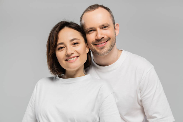 cheerful husband and tattooed wife with short brunette hair standing together in white t-shirts and looking at camera isolated on grey background in studio, happy couple  - Zdjęcie, obraz