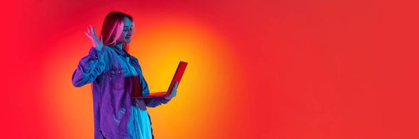 Banner with young girl, student wearing headphones and holding laptop looking at screen and smiling over red background in neon light. Concept of emotions, student life, youth, study, online education - Fotografie, Obrázek