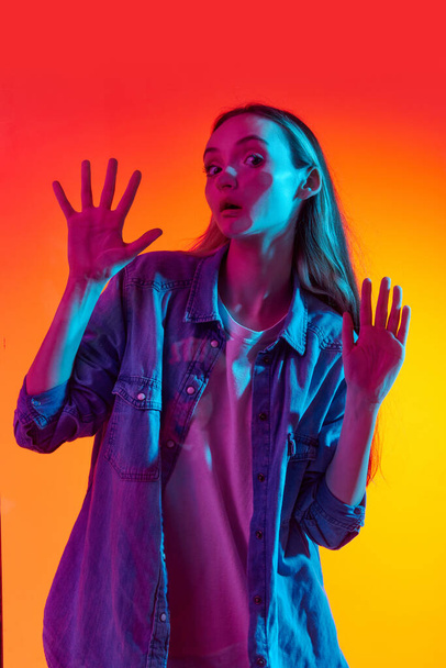 Funny meme. Portrait of one young, adorable girl, teenager looking at camera through glass with shocked face over red studio background in neon light. Concept of human emotions, fun, ad - Фото, зображення