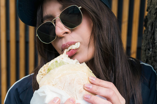Νεαρή μελαχρινή κοπέλα σε casual κοστούμι ρούχα και γυαλιά ηλίου τρώει, δαγκώνει fast food burger εξωτερική στο δρόμο. Φωτογραφία από το κεφάλι, κοντινό πορτραίτο. - Φωτογραφία, εικόνα