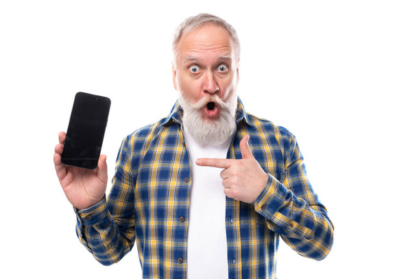 sorpreso anziano pensionato uomo dai capelli grigi con la barba in una camicia che tiene uno smartphone su uno sfondo bianco. - Foto, immagini