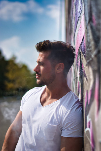 Homem muscular atraente apoiando-se na parede de grafite colorido, vestindo t-shirt branca - Foto, Imagem