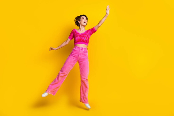 Full length photo of impressed nice carefree woman wear pink knit top pants jumping look empty space isolated on yellow color background. - Zdjęcie, obraz