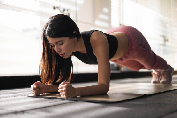 details of physical activity, performing a plank to strengthen body a slim woman with long latin hair wears sportswear, healthy lifestyle - Photo, Image