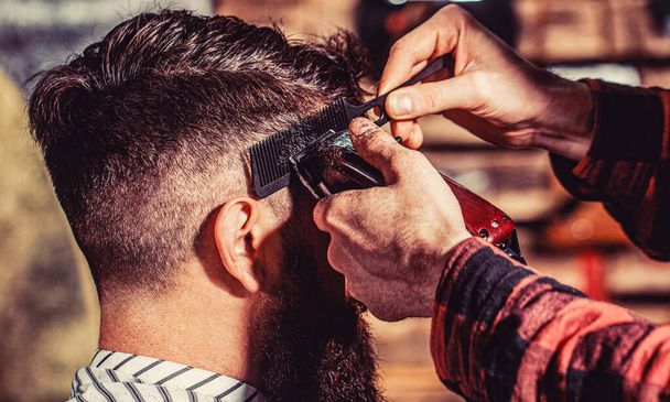 Peluquero trabaja con el cortador de pelo. Hipster cliente cortarse el pelo. Manos de peluquero con cortapelos, de cerca. Barbudo en la barbería. Hombre visitando peluquero en barbería. - Foto, Imagen