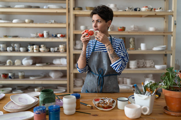 Une jeune artisanale française créative réfléchie se repose sur le lieu de travail dans un atelier de poterie, debout à la table de travail, buvant du thé et regardant de côté. Femme céramiste dans tablier prendre une pause dans l'atelier de céramique - Photo, image