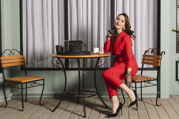 Elegant brunette lady with curly hair in red clothes, polka dot sheer lace gloves is sits in a pavement cafe, drinking coffee with a donut. - Фото, изображение