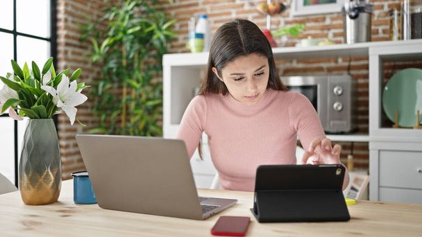 Young beautiful hispanic woman using laptop and touchpad sitting on table at dinning room - 写真・画像