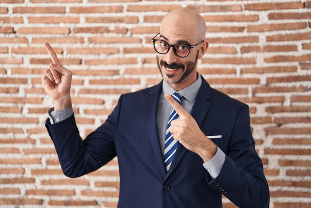 Bald man with beard wearing business clothes and glasses smiling and looking at the camera pointing with two hands and fingers to the side.  - Photo, Image