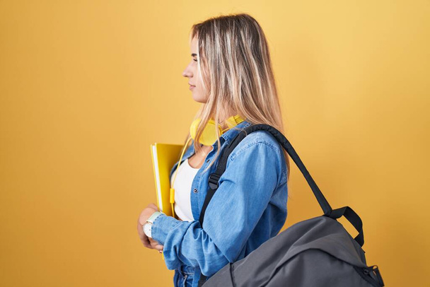 Junge blonde Frau trägt Studentenrucksack und hält Bücher zur Seite, entspannte Profil-Pose mit natürlichem Gesicht und selbstbewusstem Lächeln.  - Foto, Bild
