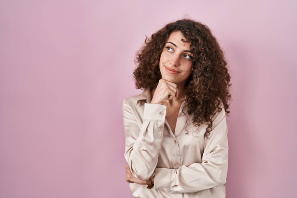 Hispanic woman with curly hair standing over pink background with hand on chin thinking about question, pensive expression. smiling with thoughtful face. doubt concept.  - Фото, изображение
