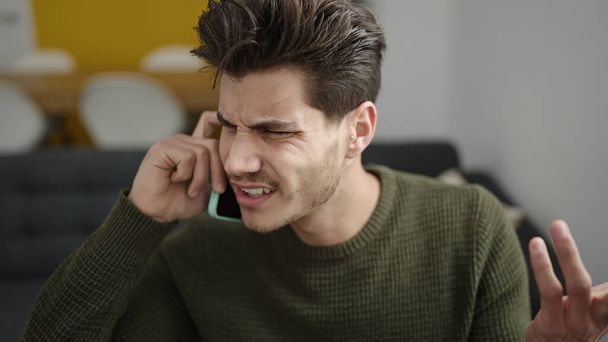 Молодий іспаномовний чоловік розмовляє по смартфону з нещасним виразом вдома
 - Фото, зображення