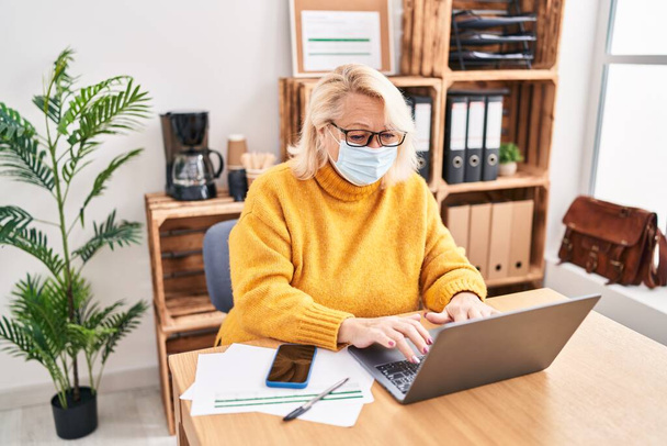 Blonde Geschäftsfrau mittleren Alters mit medizinischer Maske arbeitet im Büro - Foto, Bild