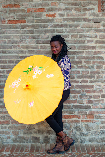Portrait of an African woman with dreadlocks braid with umbrella parasol. Happy young woman feeling confident in her style. Fashionable woman standing in the street of old village against stone wall. - Photo, Image