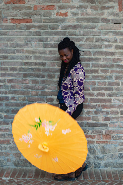 Portré egy afrikai nőről, aki napernyővel van befonva. Boldog fiatal nő, magabiztos a stílusában. Divatos nő áll az utcán az öreg falu kőfal. - Fotó, kép