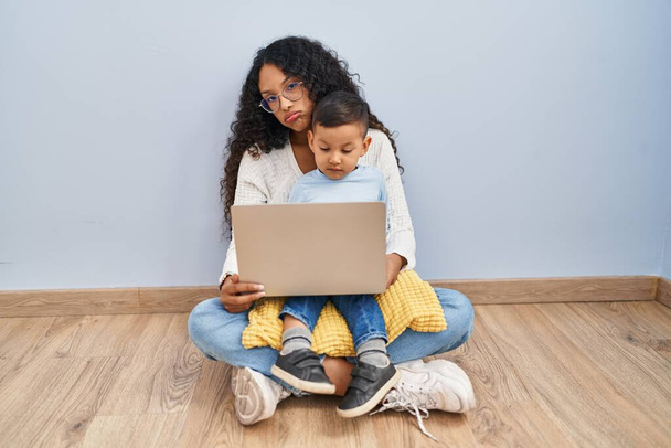 Young hispanic mother and kid using computer laptop sitting on the floor looking sleepy and tired, exhausted for fatigue and hangover, lazy eyes in the morning.  - Photo, Image