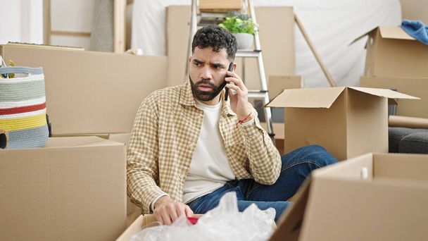 Young hispanic man talking on smartphone unpacking cardboard box at new home - Photo, Image