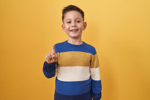 Mały latynoski chłopiec stojący nad żółtym tłem uśmiechający się z pomysłem lub pytaniem wskazującym palcem na szczęśliwą twarz, numer jeden  - Zdjęcie, obraz