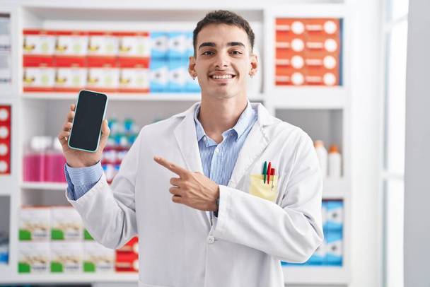 Handsome hispanic man working at pharmacy drugstore showing smartphone screen smiling happy pointing with hand and finger  - Фото, изображение