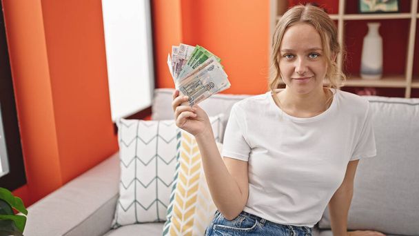 Nuori blondi nainen hymyilee luottavainen tilalla Venäjän ruplaa seteleitä kotona - Valokuva, kuva