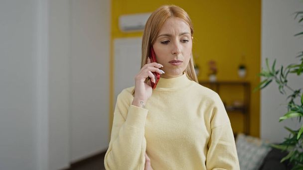 Young blonde woman talking on smartphone with serious expression at home - Fotoğraf, Görsel