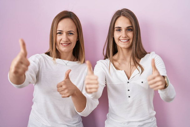 Middle age mother and young daughter standing over pink background approving doing positive gesture with hand, thumbs up smiling and happy for success. winner gesture.  - Фото, изображение