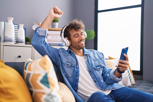Young hispanic man dancing and listening to music sitting on sofa at home - Photo, Image