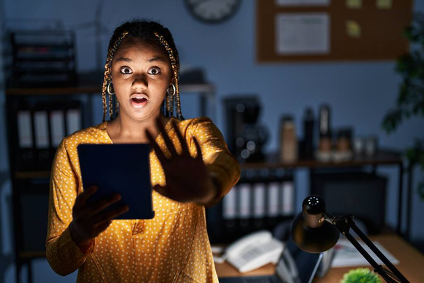 Donna afroamericana con trecce che lavora in ufficio di notte con tablet facendo gesto di stop con palmi delle mani, espressione arrabbiata e frustrazione  - Foto, immagini