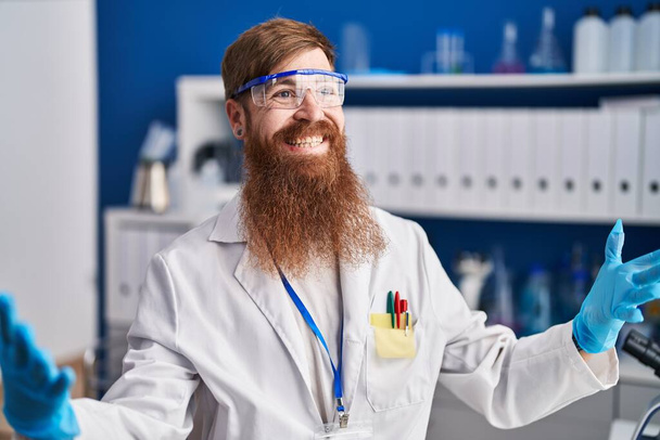 Fiatal vörös hajú férfi tudós mosolygós magabiztos beszél a laboratóriumban - Fotó, kép