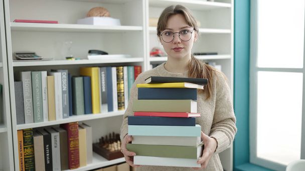 Jeune étudiante blonde tenant des livres avec une expression détendue à la bibliothèque de l'université - Photo, image