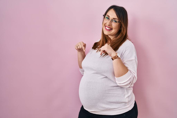 Terhes nő áll a rózsaszín háttér néz magabiztos mosollyal az arcon, mutatva magát ujjaival büszke és boldog.  - Fotó, kép