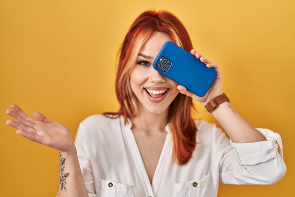 Mujer caucásica joven que cubre el ojo con un teléfono inteligente celebrando el logro con sonrisa feliz y expresión ganadora con la mano levantada  - Foto, Imagen