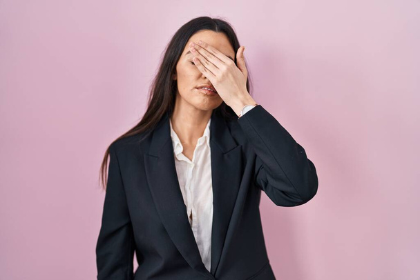 Young brunette woman wearing business style over pink background covering eyes with hand, looking serious and sad. sightless, hiding and rejection concept  - Fotografie, Obrázek
