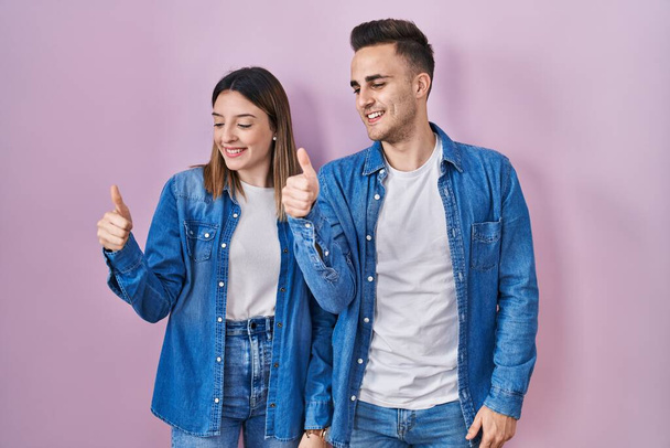 Fiatal spanyol pár áll a rózsaszín háttér felett, büszkén néznek, mosolyognak, és felemelik a hüvelykujjukat oldalra.  - Fotó, kép