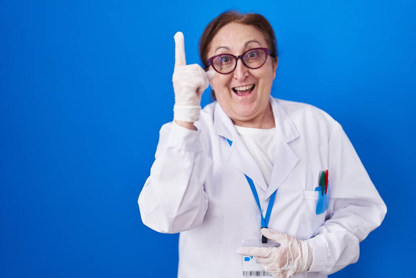 Senior woman with glasses wearing scientist uniform pointing finger up with successful idea. exited and happy. number one.  - Valokuva, kuva