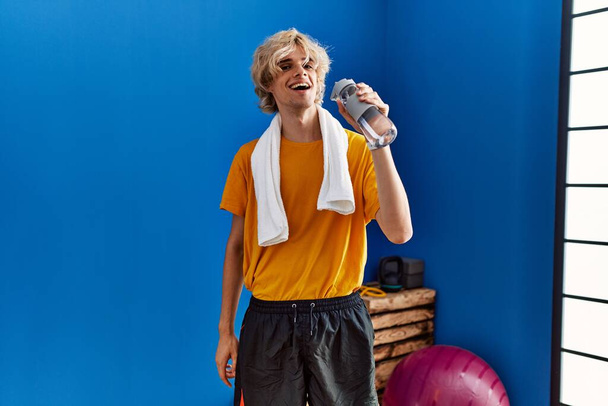 Νεαρός ξανθός άνδρας χαμογελά αυτοπεποίθηση πόσιμο νερό στο αθλητικό κέντρο - Φωτογραφία, εικόνα