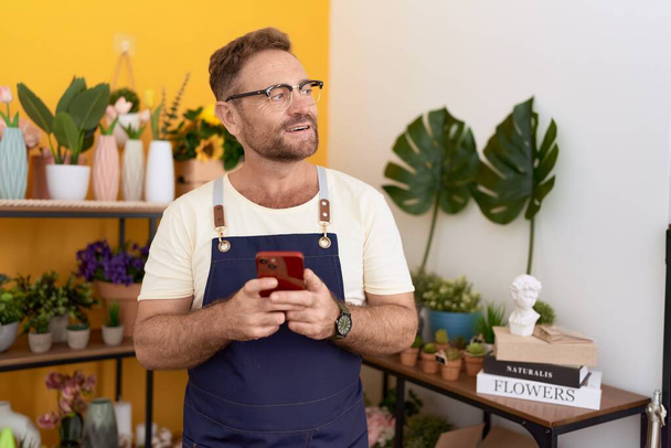 Középkorú férfi virágárus mosolygós magabiztos segítségével smartphone virágüzlet - Fotó, kép
